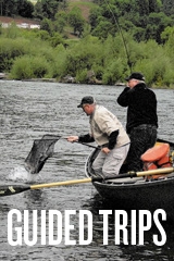 Two men in a row boat, one with a reel with a fish on the line, and another with a net trying to land the fish
